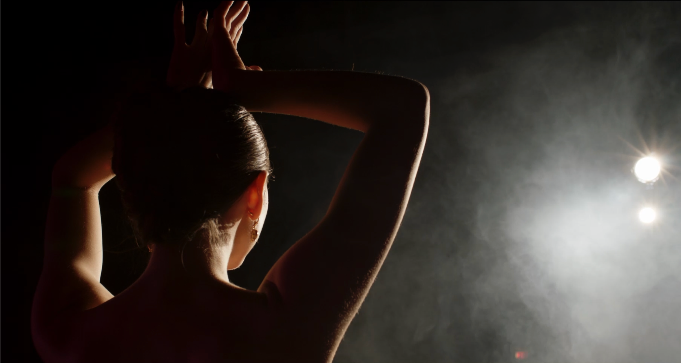 Back view of a female dancer with her arms raised and hands on her head, silhouette illuminated by bright stage lights.