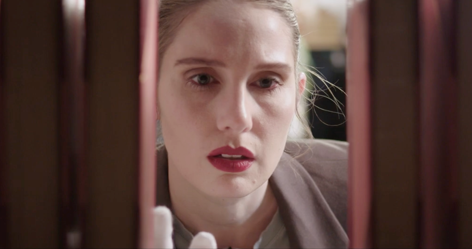 A woman with red lipstick and blonde hair looks through the gap between two books on a bookshelf.