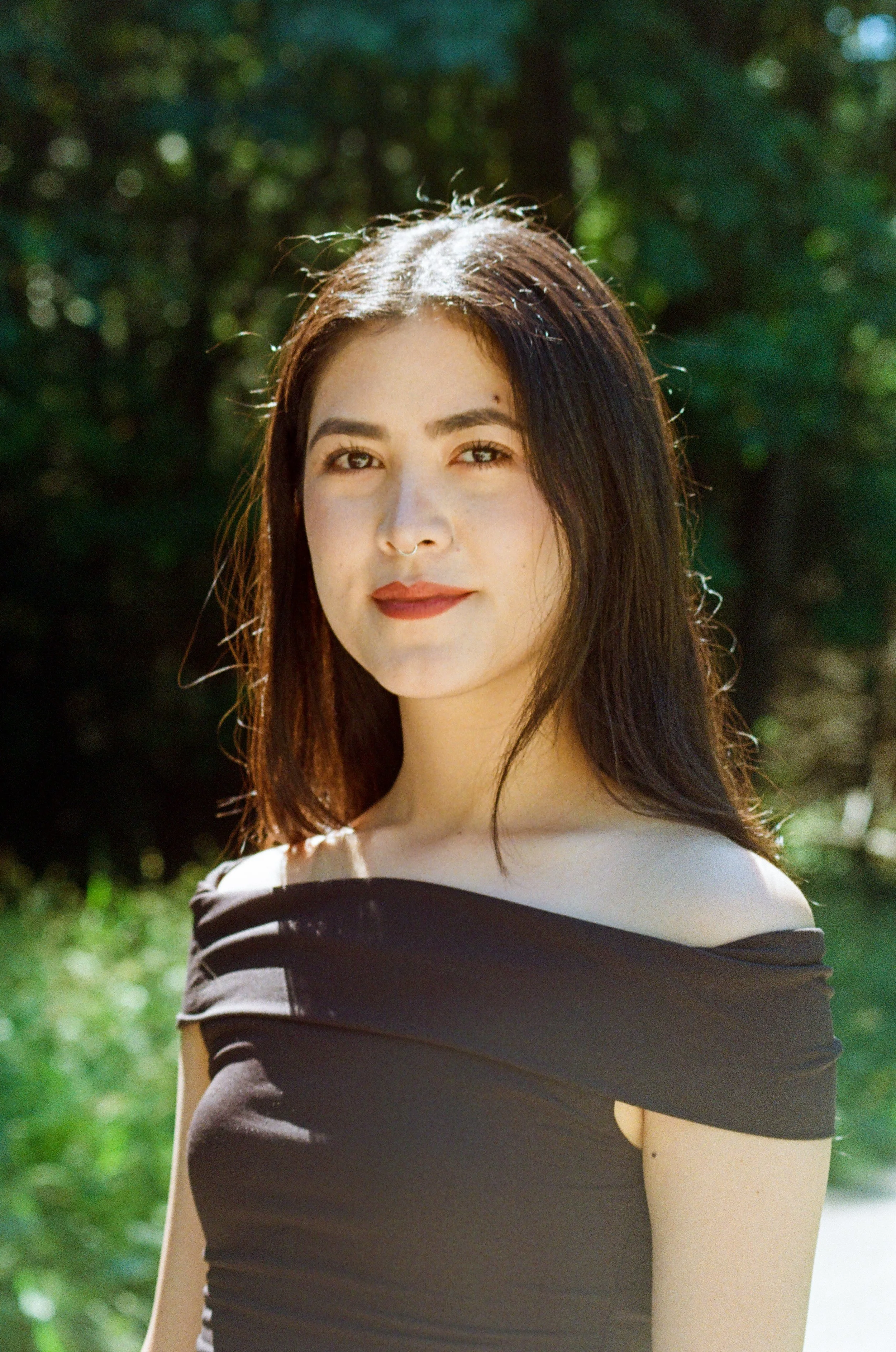 A woman with long brown hair, wearing a black off-the-shoulder top, with green blurred foliage in the background.