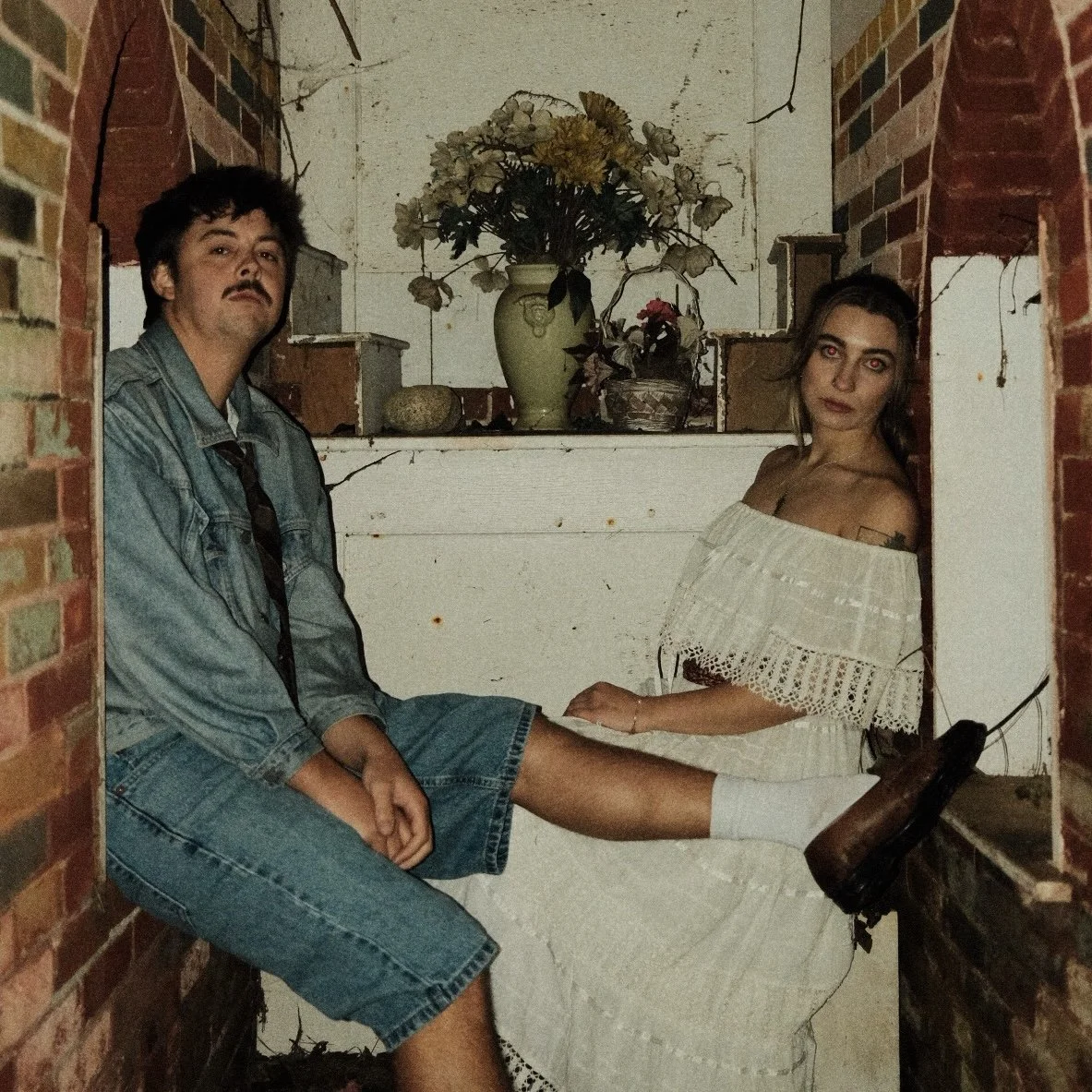 A man and woman sitting in a small, vintage space with brick walls and white wooden panels, decorated with a flower vase and potted plants.