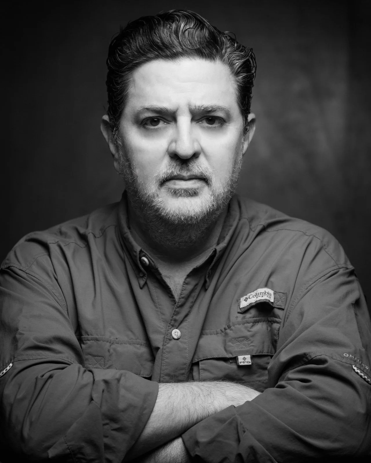 Retrato en blanco y negro de un hombre con barba y cabello oscuro, con expresión seria, usando una chaqueta Columbia y con los brazos cruzados.