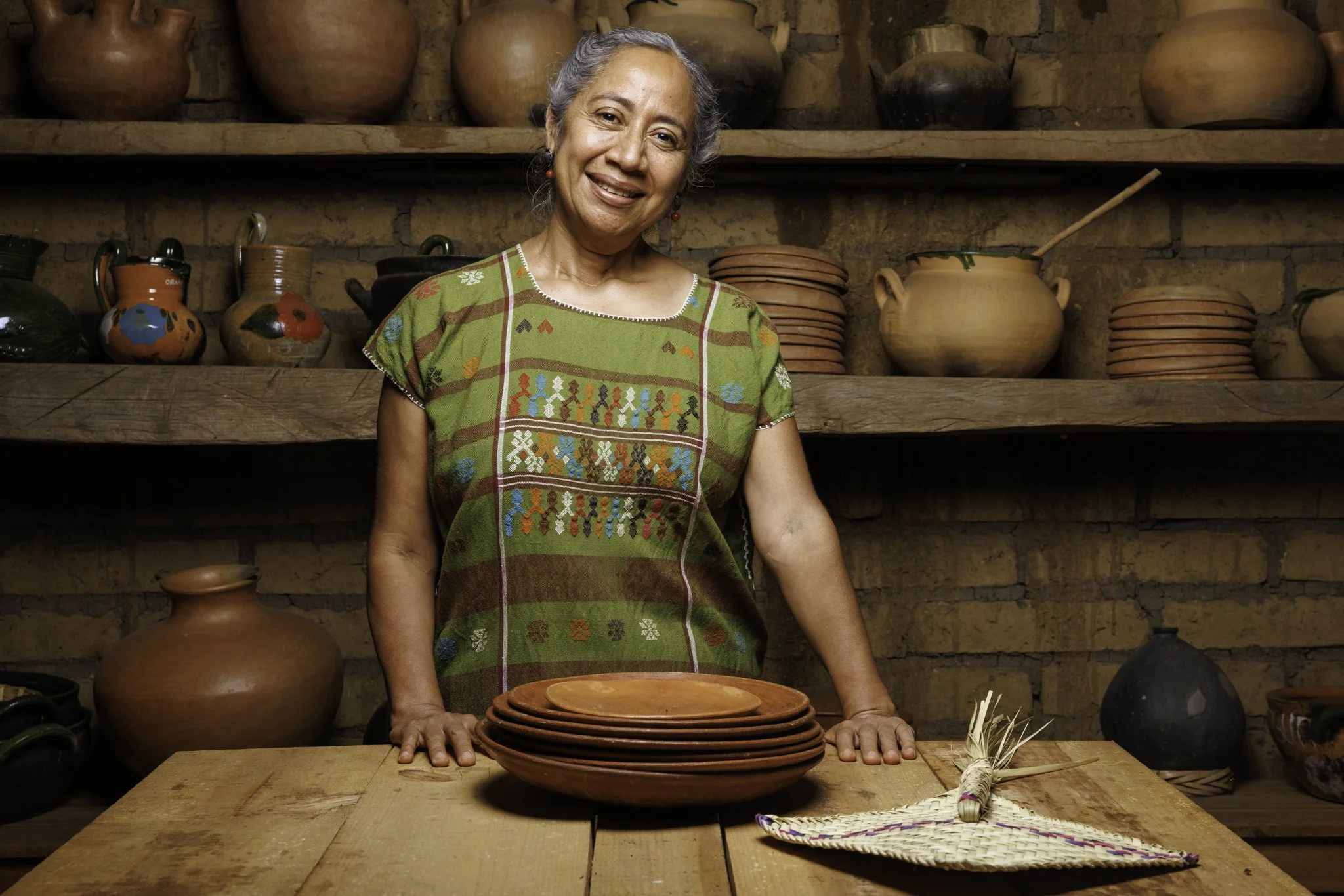 Mujer mayor sonriente con ropas tradicionales mexicanas, rodeada de cerámicas y utensilios de barro en un ambiente rústico.