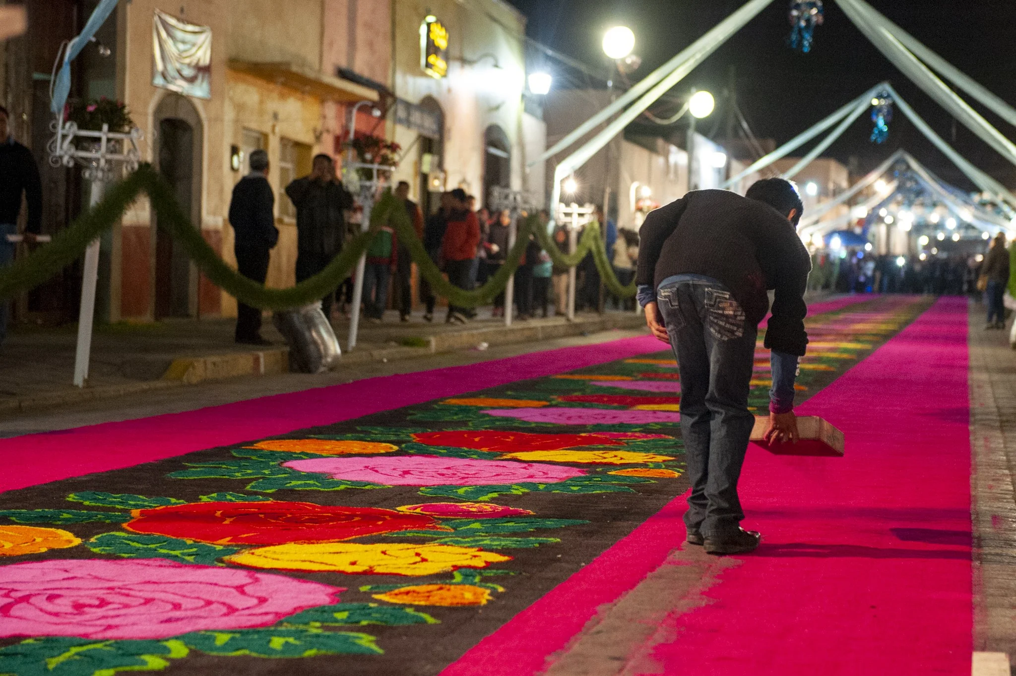 Personas trabajando en la elaboración de un tapete colorido en la calle durante la noche, con decoraciones y luces en el entorno urbano.