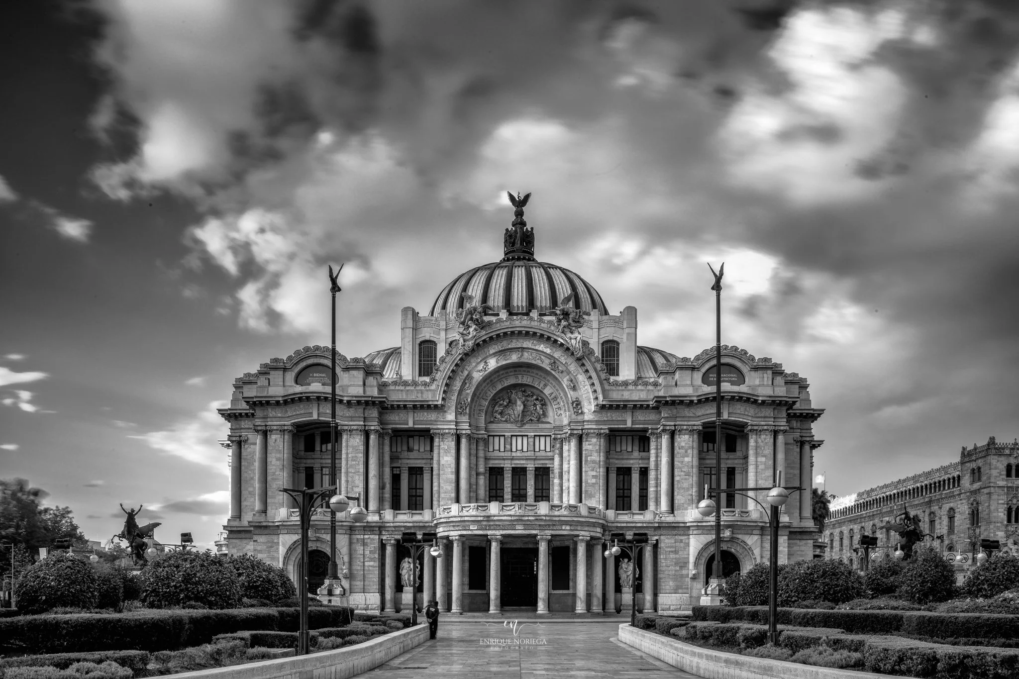 Edificio histórico con cúpula y adornos arquitectónicos, en un entorno urbano con árboles y farolas, imagen en blanco y negro.