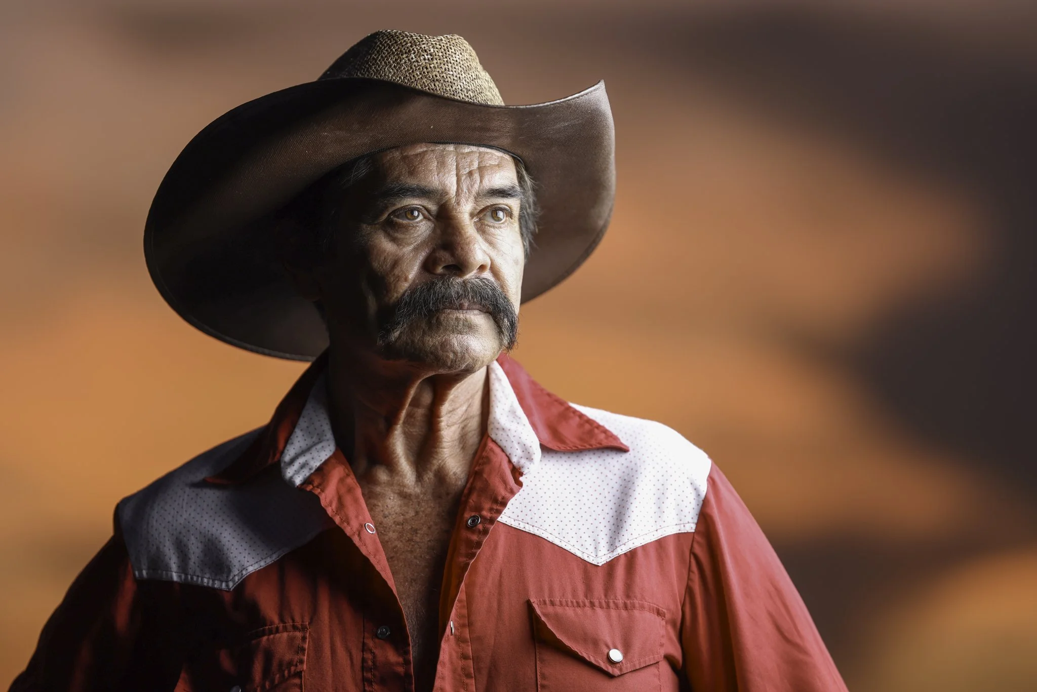 Hombre con sombrero de vaquero, barba y bigote, vestido con camisa de vestir roja y chaqueta en tonos oscuros, frente a un fondo difuso en tonos naranjas y marrones.