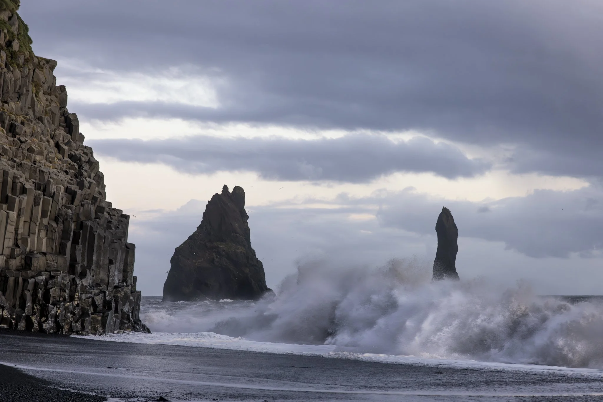 Escena de una costa con rocas en el mar y olas rompiendo contra ellas, con formaciones rocosas altas en el fondo y un cielo nublado.