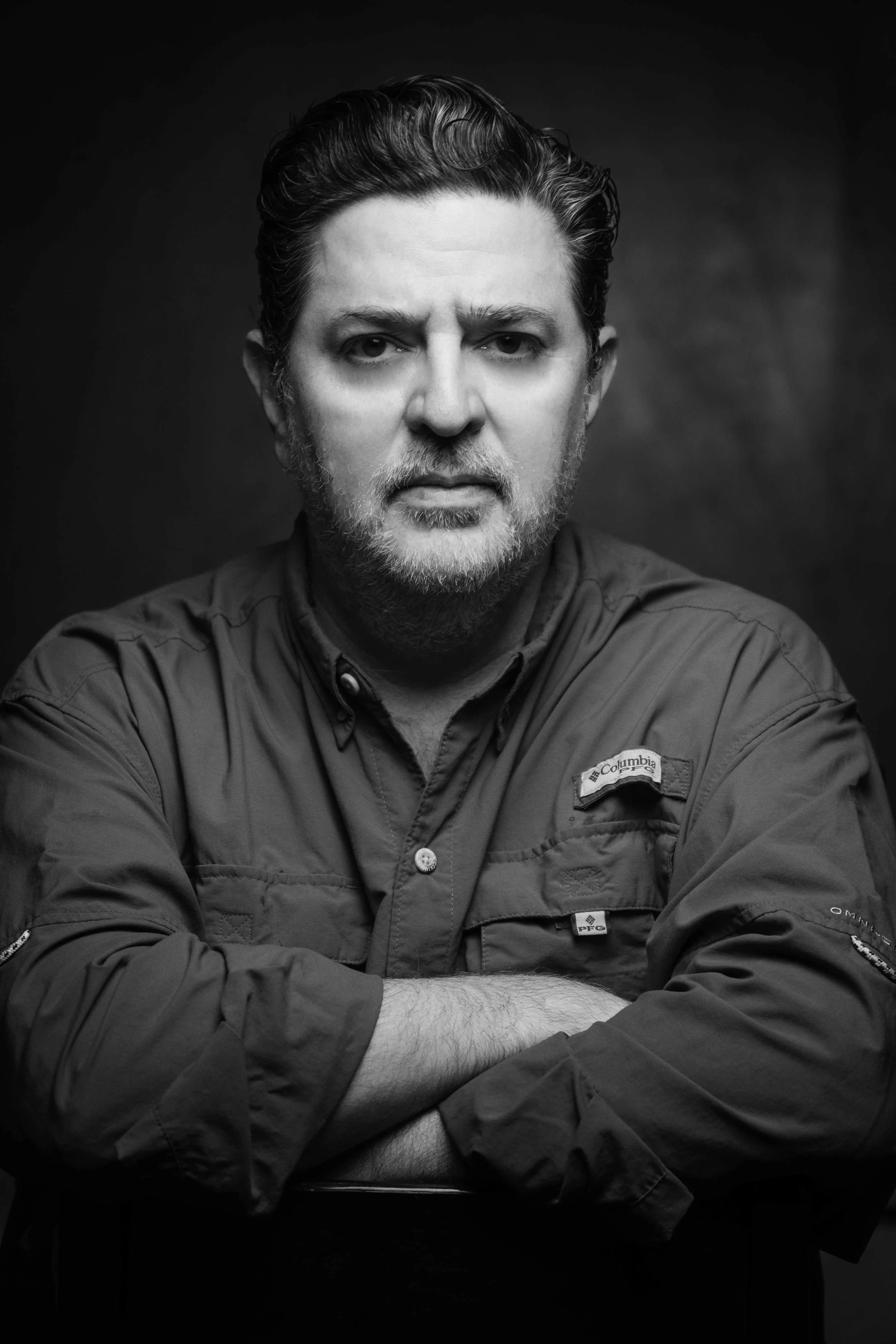Retrato en blanco y negro de un hombre con cabello y barba canosos, usando una chaqueta de outdoors con parche de Columbia, con expresión seria y brazos cruzados, en un fondo oscuro.