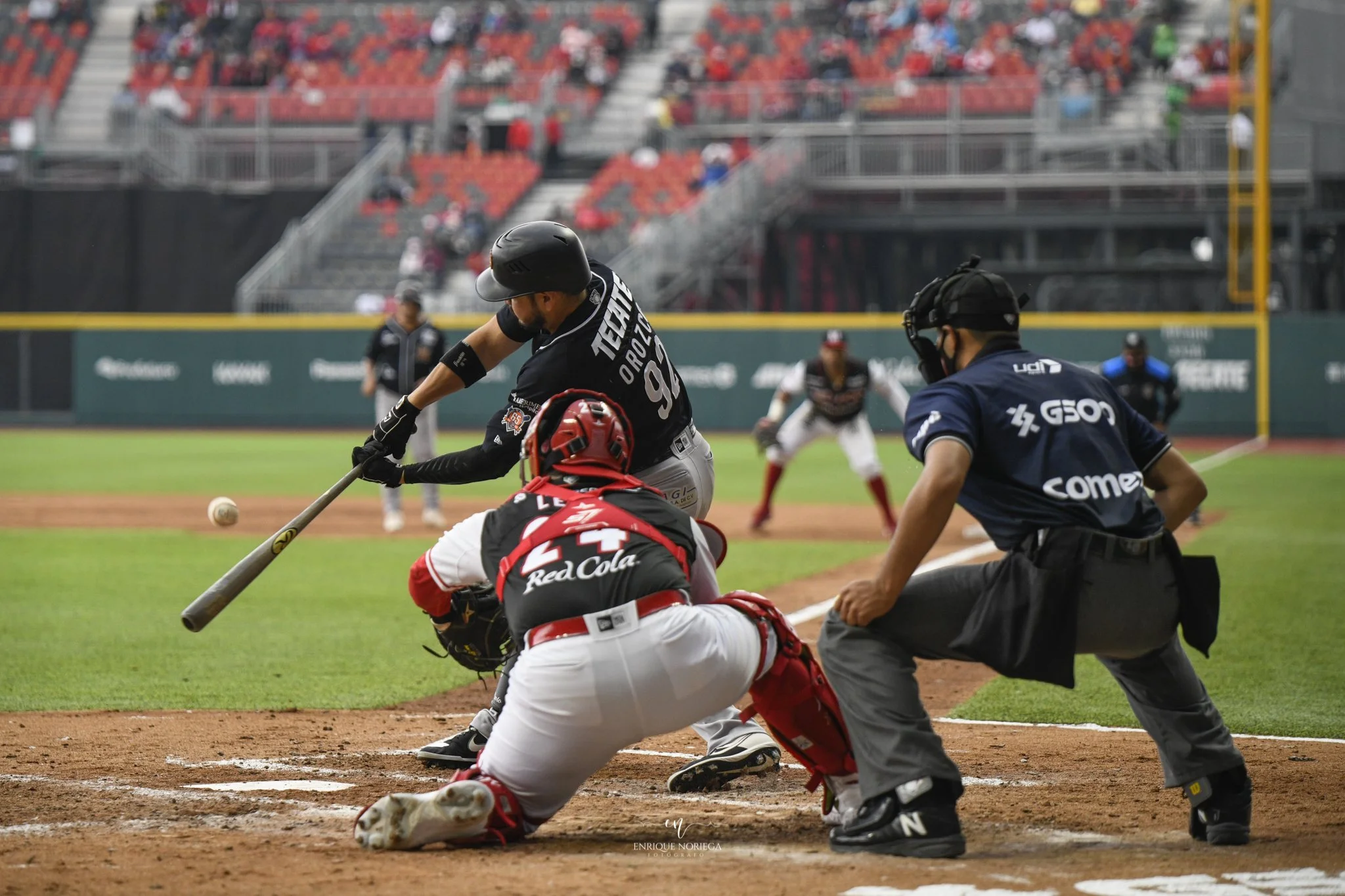 Jugadores de béisbol en acción en un campo, con un pitcher lanzando la pelota y un receptor en posición de atraparla, en un estadio con espectadores