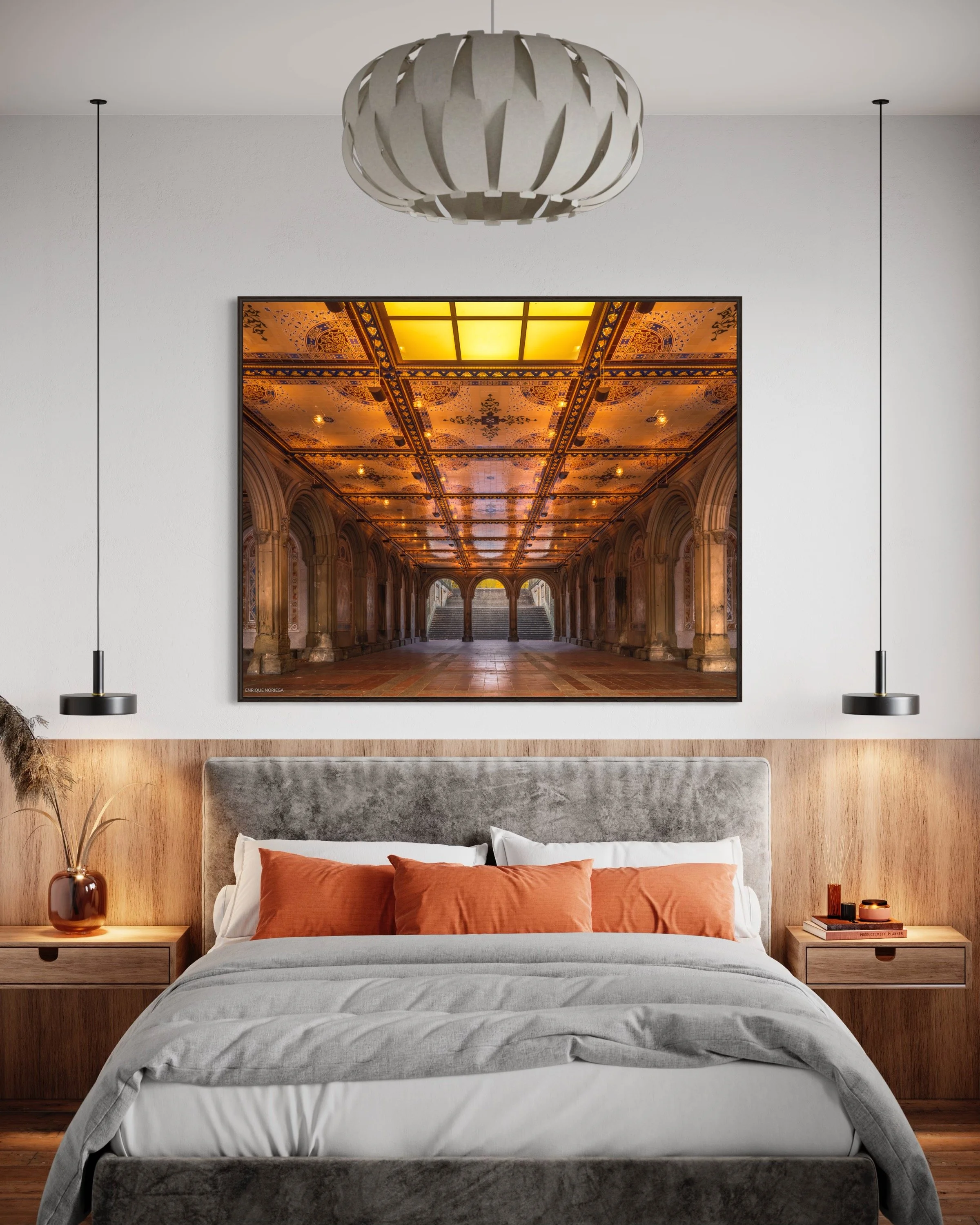 Cama con ropa blanca y cojines naranjas, dos mesas de noche de madera a cada lado, lámparas colgantes negras y cuadro de un pasillo con techo dorado y arcos.