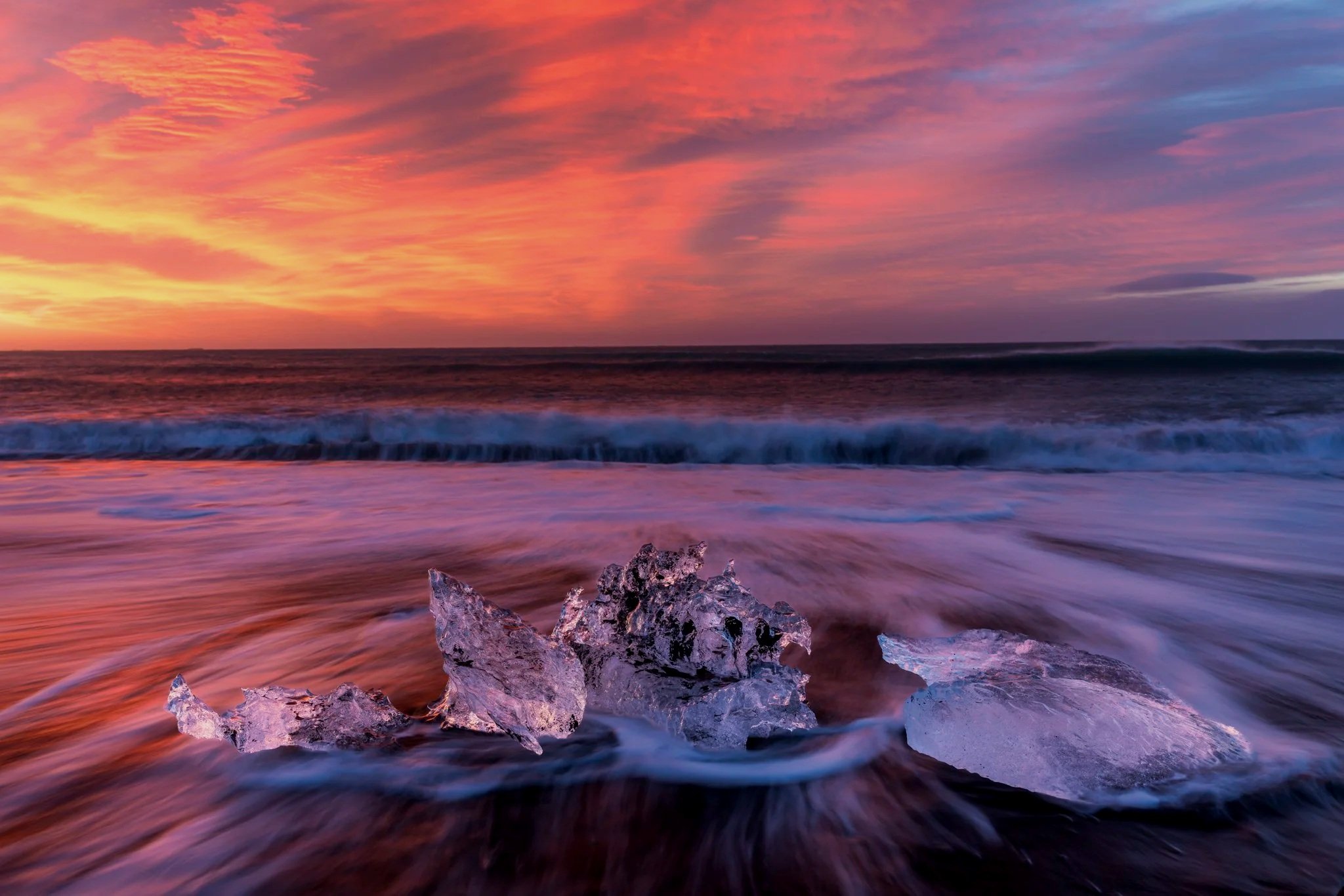 Atardecer en la playa con olas suaves y cristales de hielo en la orilla, cielo con nubes color rosa, naranja y morado.