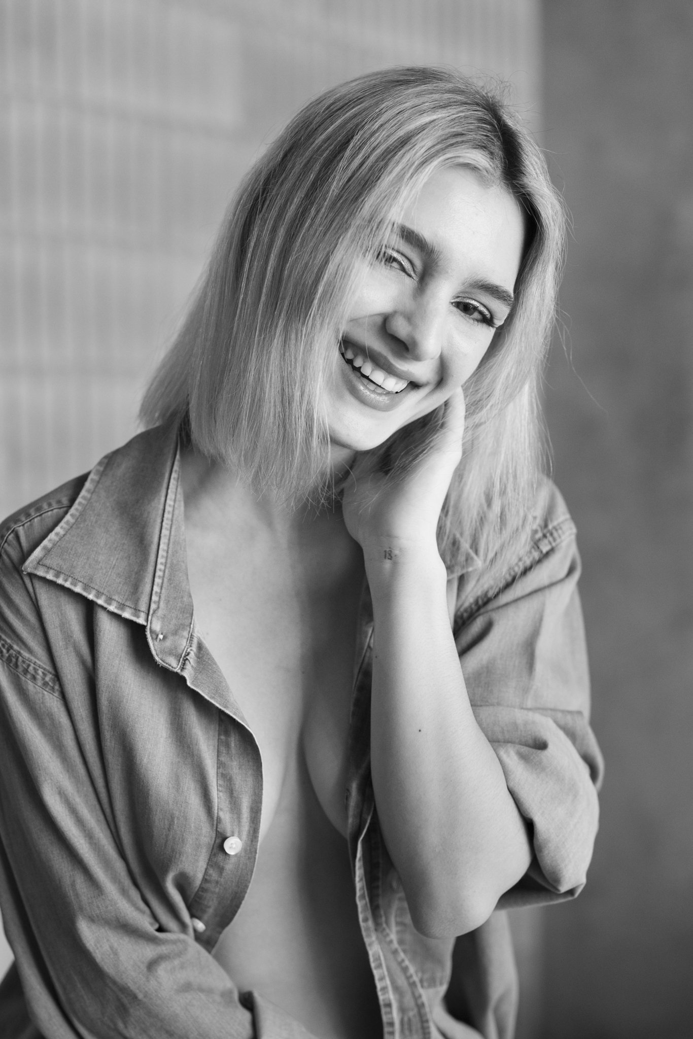 Retrato en blanco y negro de una mujer sonriendo y tocándose la cara, con cabello rubio, usa una camisa de mezclilla