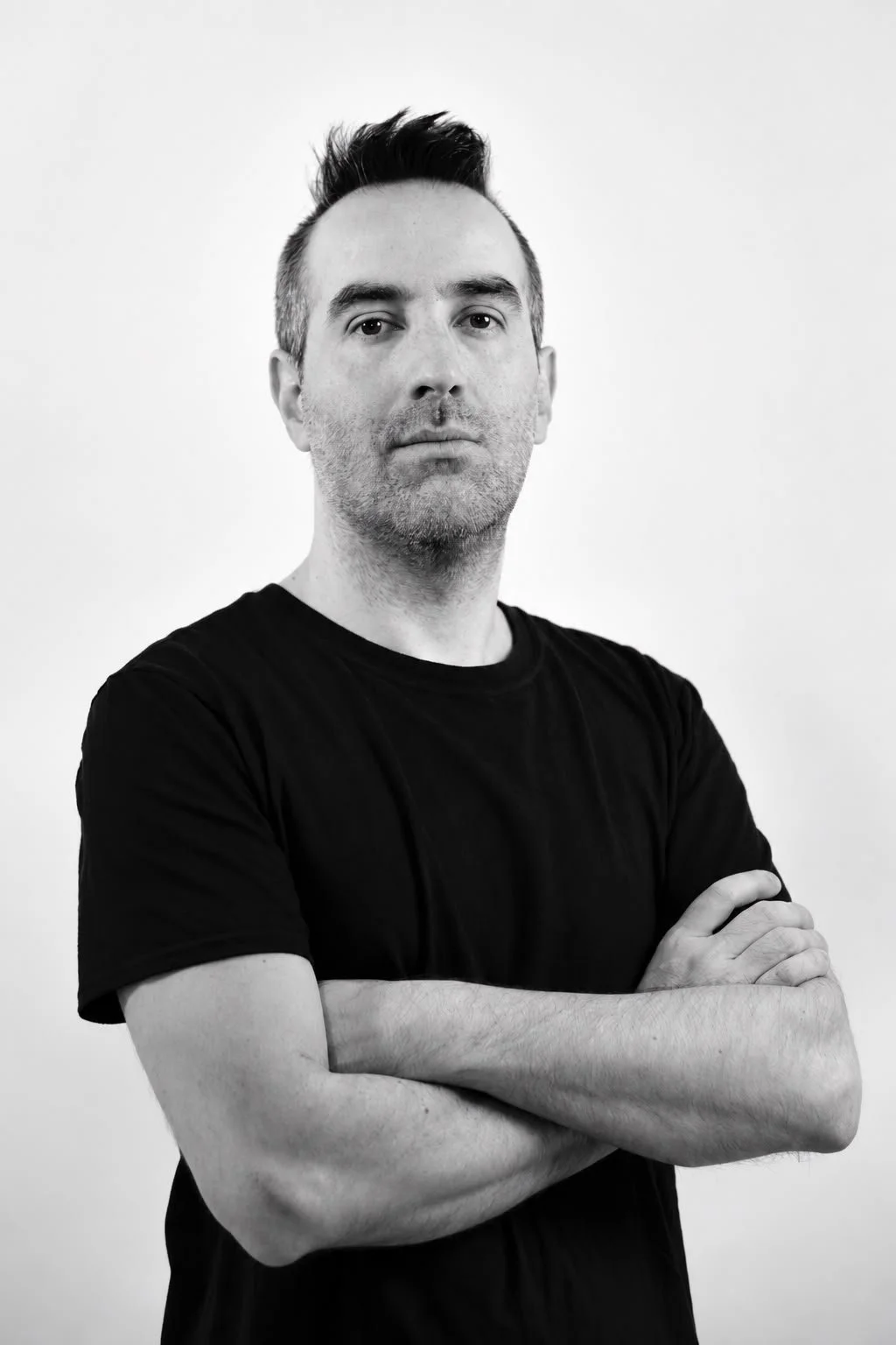 Primer plano de un hombre con cabello oscuro y despeinado, usando una camiseta negra, con los brazos cruzados y expresión seria, en un fondo blanco.