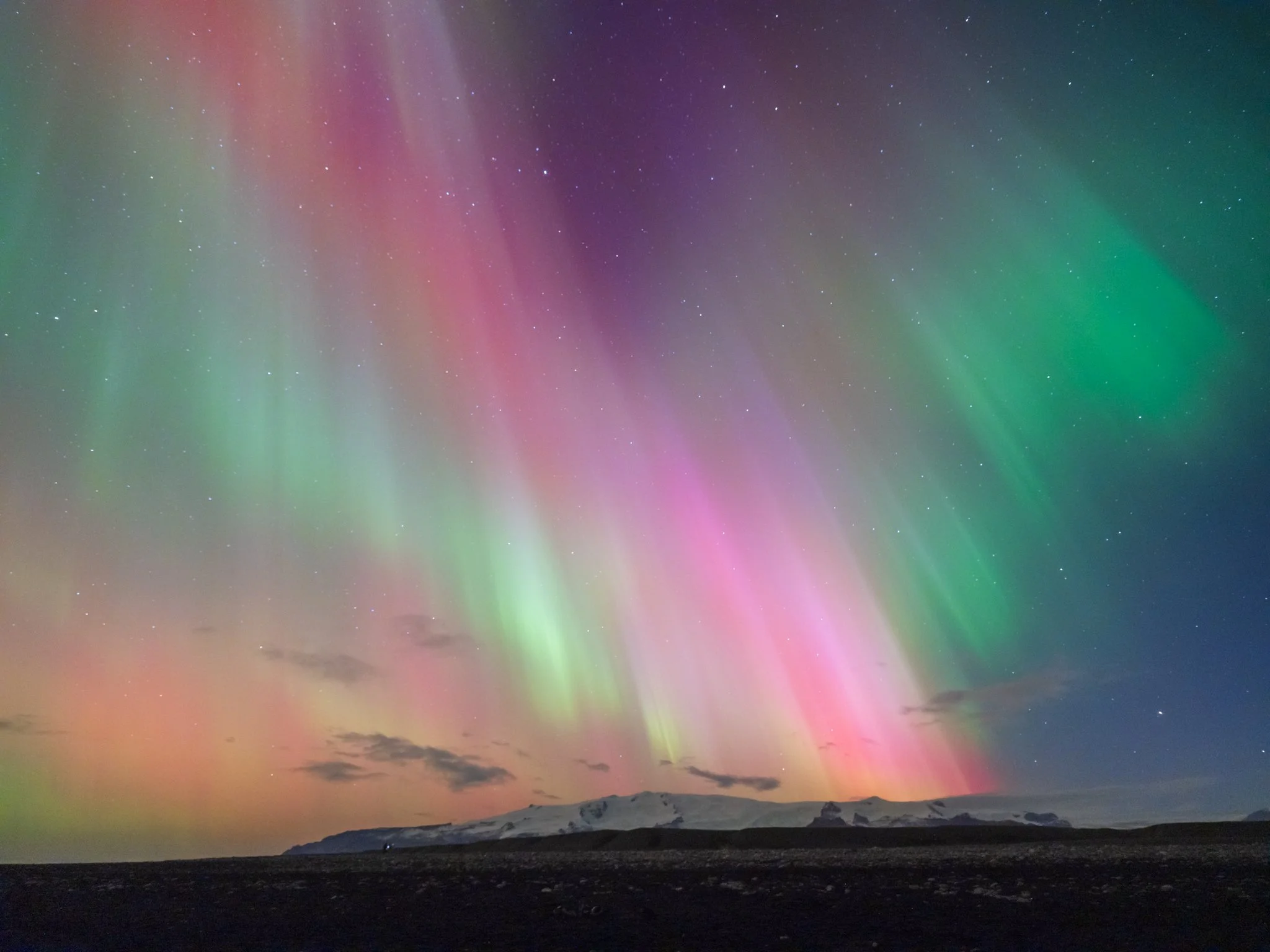 Auroras boreales de colores rosa, verde y púrpura en el cielo nocturno sobre montañas y tierra oscura.