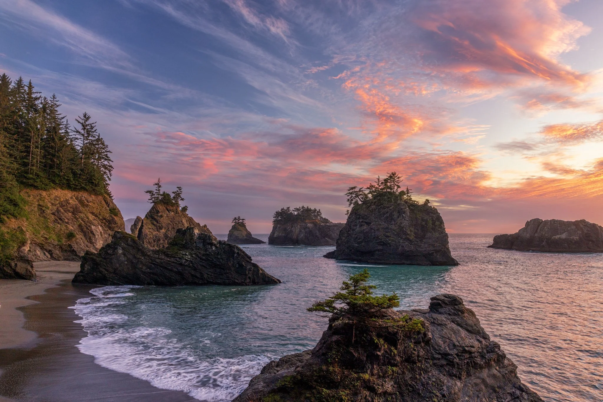 Costa rocosa con árboles y rocas en el mar durante el atardecer, con nubes de colores rosados y anaranjados en el cielo.