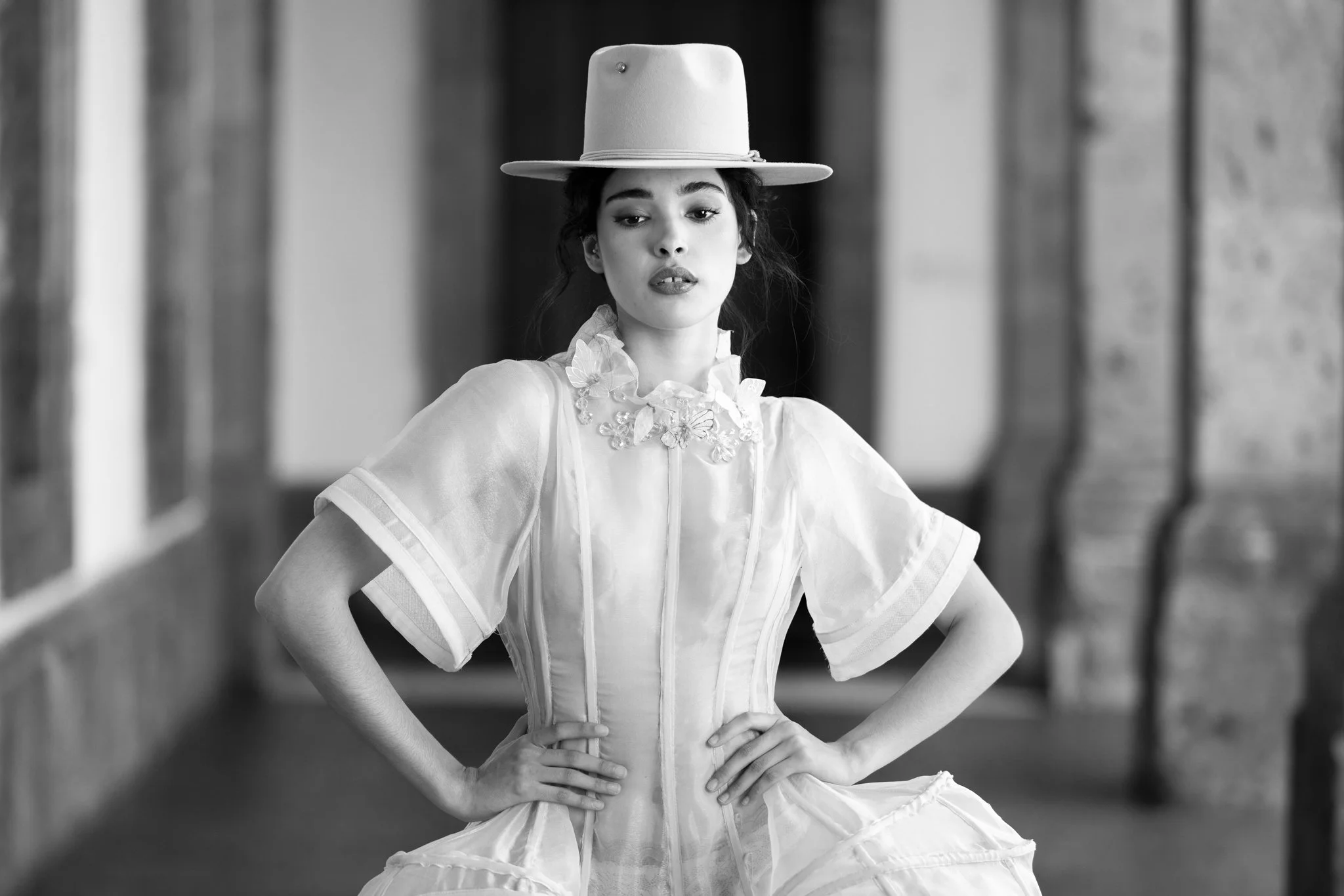 Mujer con sombrero grande y vestido elegante, posando con las manos en las caderas en un entorno urbano, en blanco y negro.
