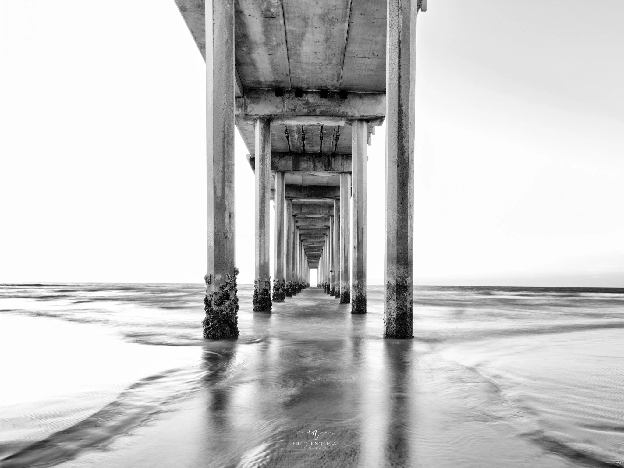 Vista desde debajo de un puente de concreto en la playa, con columnas que se extienden en línea recta hacia el horizonte, reflejándose en el agua.