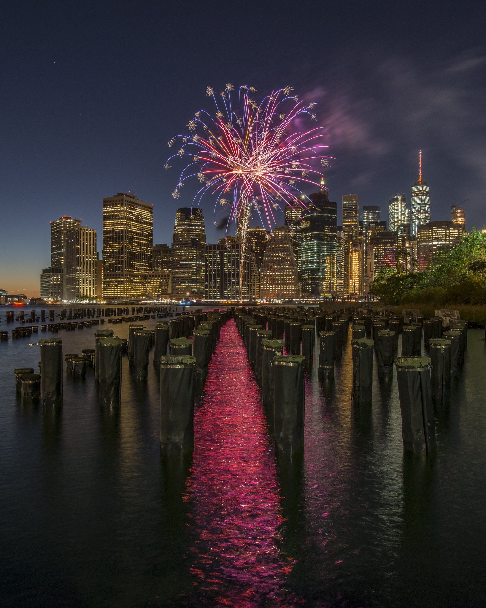 Fuegos artificiales iluminan el cielo nocturno sobre un horizonte de edificios de una ciudad moderna, reflejándose en el agua de un muelle con pilares de madera.