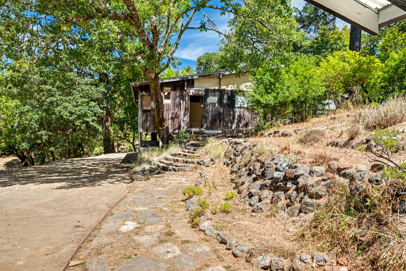A rustic wooden house on a hillside, surrounded by lush green trees. A stone pathway leads up to the entrance with steps, and there is a rocky slope on the right side of the pathway.
