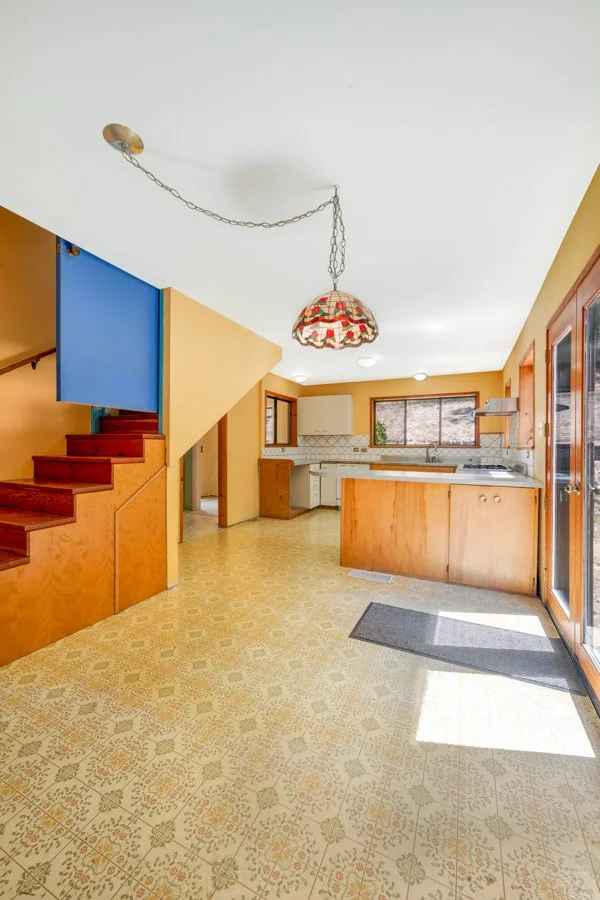 Kitchen with wooden cabinets, a tiled backsplash, a window over the sink, and a patterned linoleum floor. An open staircase with red steps leads up on the left. A stained glass hanging lamp is on the ceiling, and a sliding glass door is on the right 