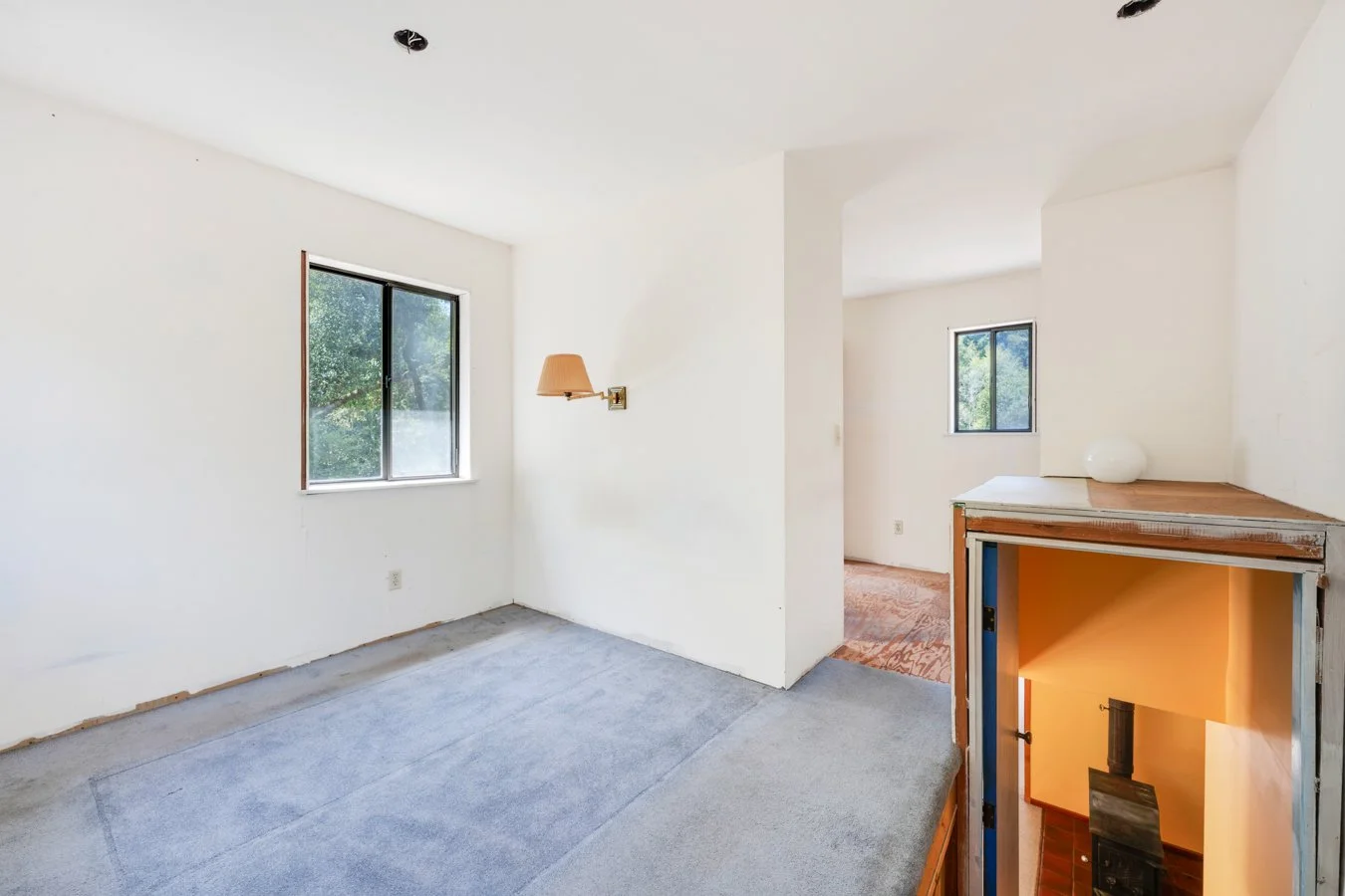 Empty living room with two windows, white walls, a wall-mounted lamp, and unfinished cabinetry with a stove, in a house under renovation.