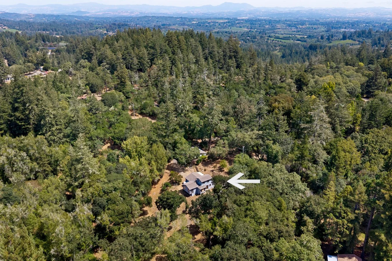 Aerial view of a forested area with a scattered residential neighborhood and a house marked with a white arrow.