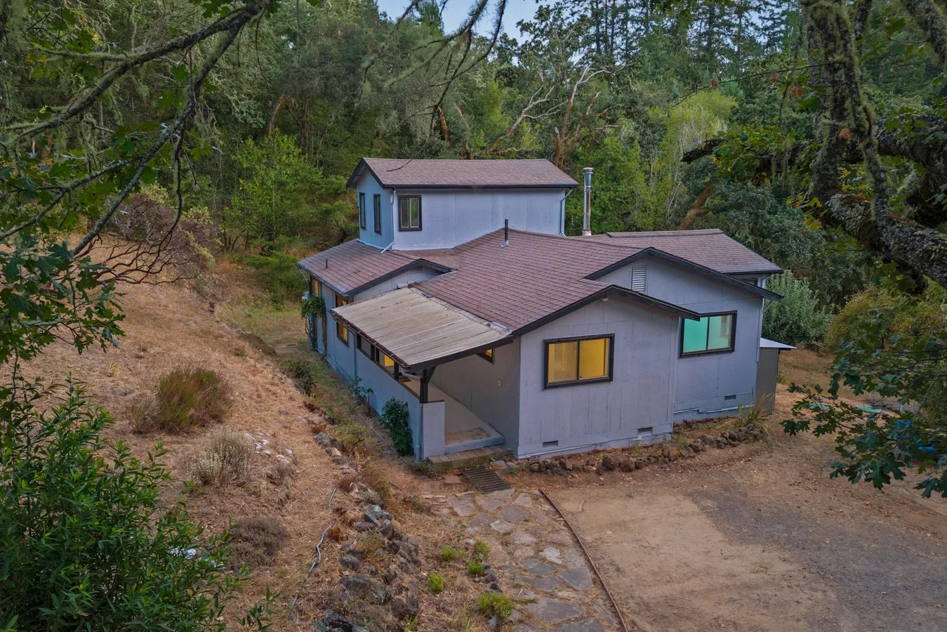 A house with multiple sections and a sloped roof, located in a wooded area with trees and a dirt path leading to the front entrance.