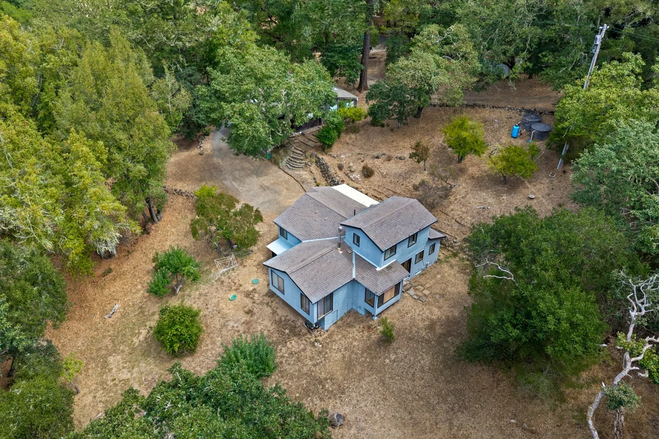 Aerial view of a two-story gray house surrounded by trees and a brown yard, with a driveway leading to the house and a small patch of lawn with some shrubs.