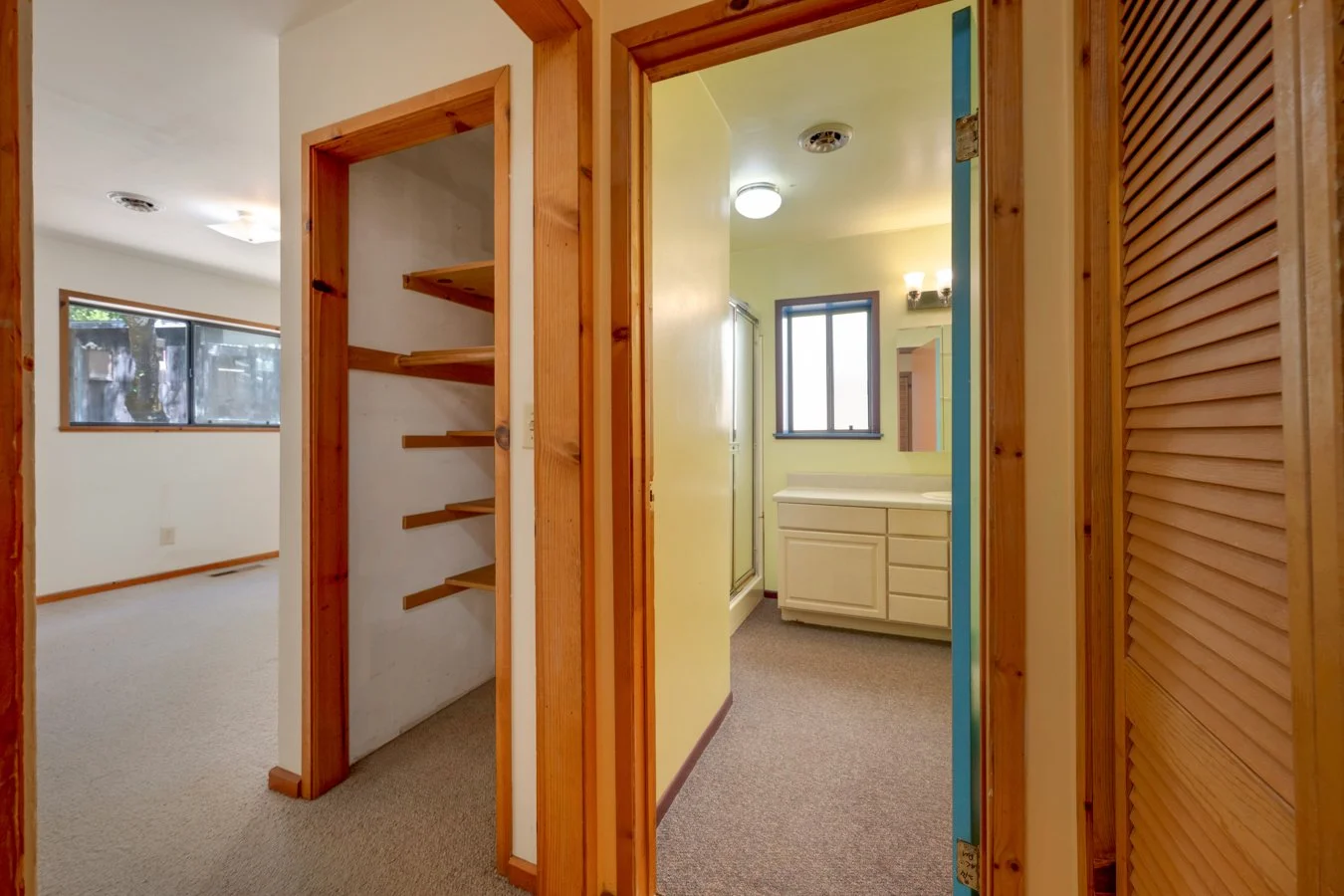 View of a small bathroom with a white vanity, a window, yellow walls, and bathroom fixtures, seen through a doorway with a wooden frame. To the left, there's a hallway with a built-in wooden shelf and a closet with wooden louvered doors.