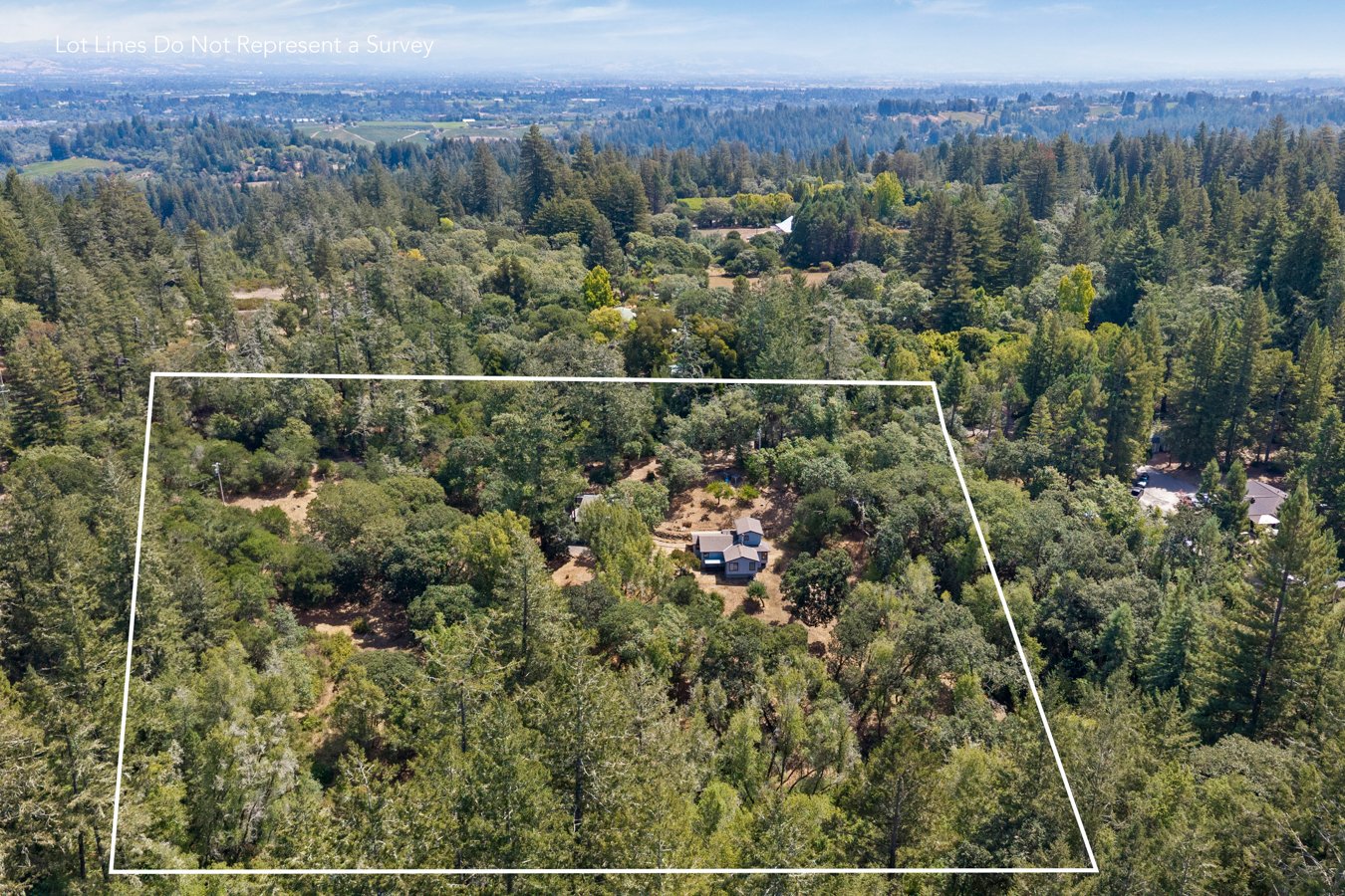 Aerial view of a wooded property outlined with white borders, featuring a small house and surrounding trees, with a distant landscape of trees and hills in the background.