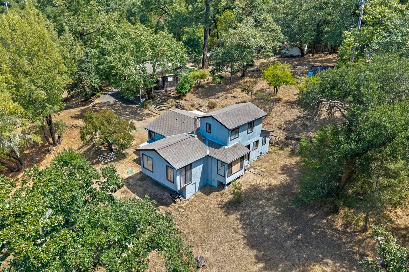An aerial view of a two-story blue house with a gable roof, surrounded by trees and a dirt yard, in a wooded area.