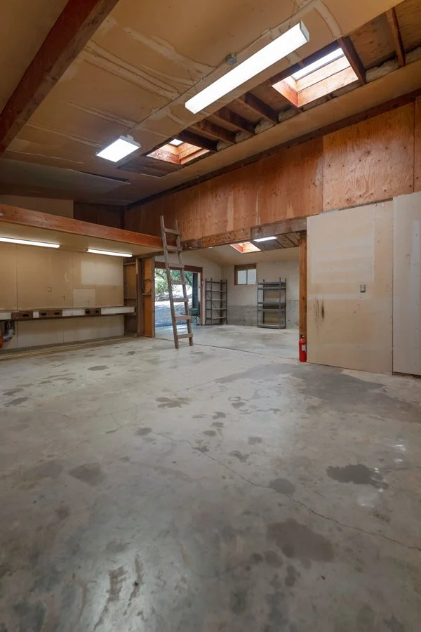 Interior of a warehouse or workshop with unfinished ceiling, skylights, fluorescent lighting, concrete floor, and shelving units.