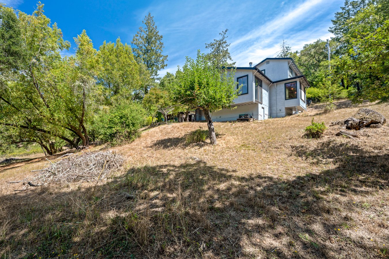 A white house with large windows on a hillside surrounded by trees and dry grass under a blue sky.
