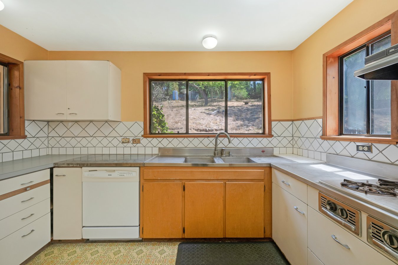 Kitchen with beige walls, wooden trim around two windows, white tile backsplash, white appliances, and a double sink facing a backyard with trees.
