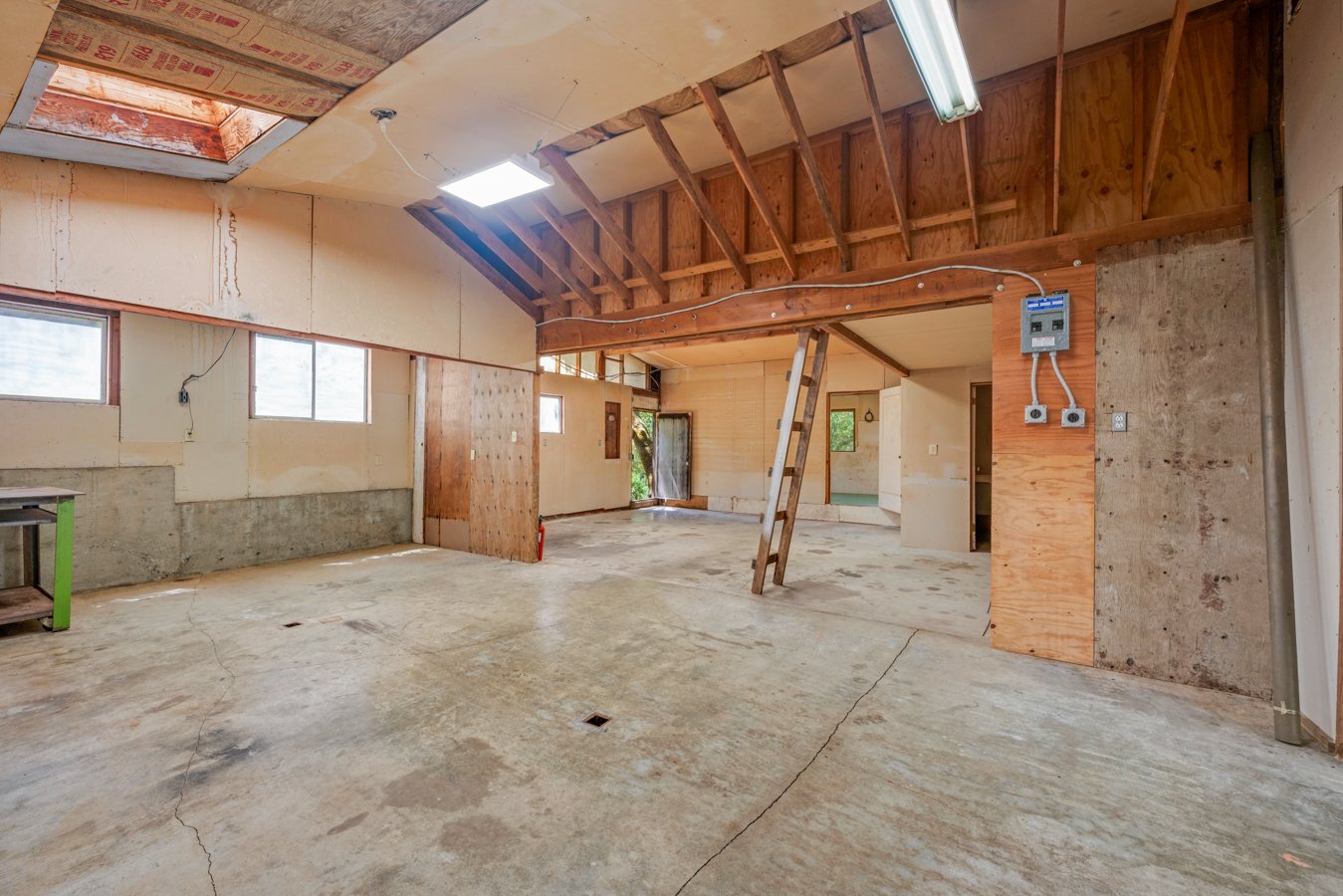 Interior of a partially renovated room with exposed wooden beams, drywall, and concrete floor. Several windows and a skylight are visible, with some construction materials and a ladder present.