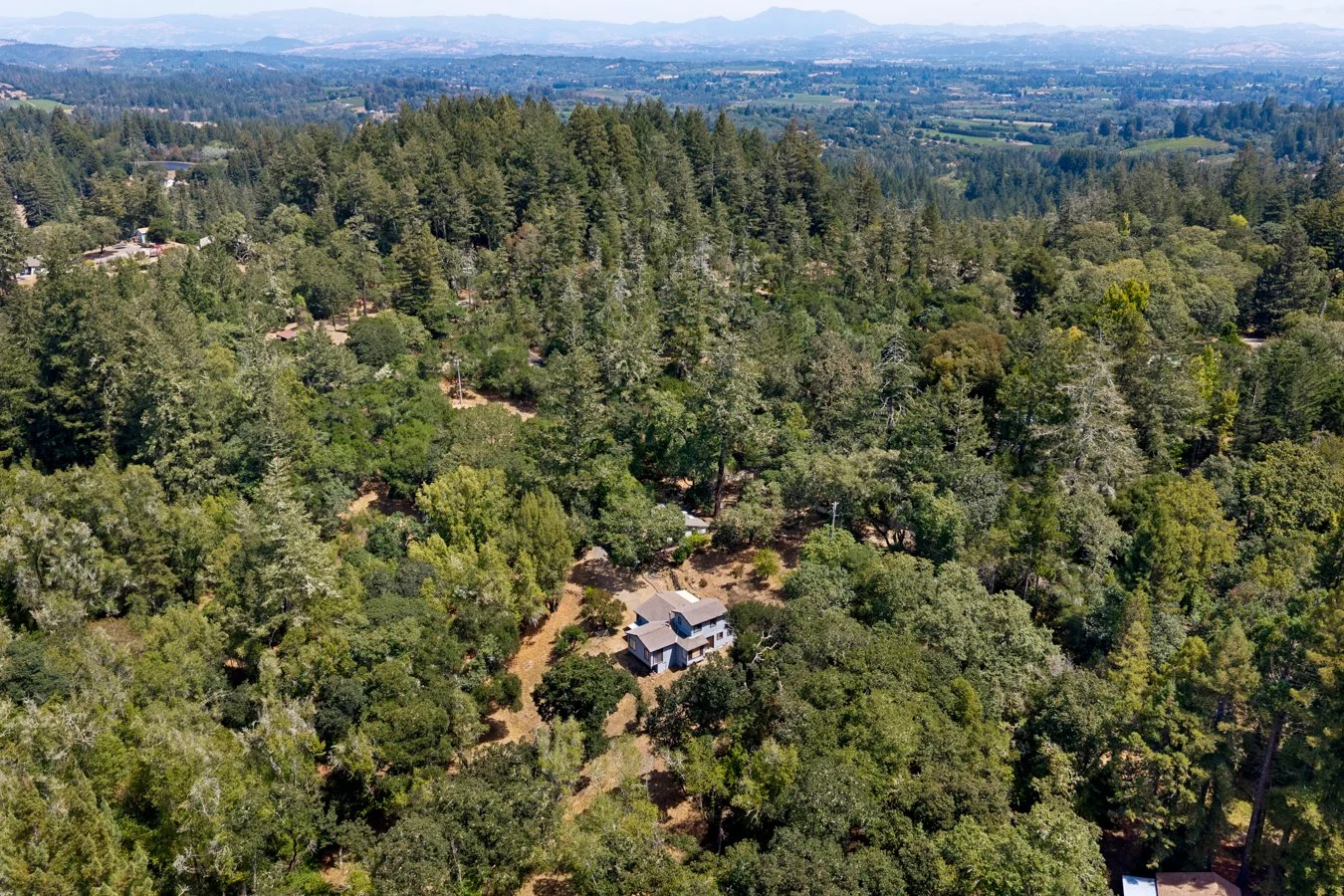 Aerial view of a house surrounded by dense forest and rolling hills in the distance.