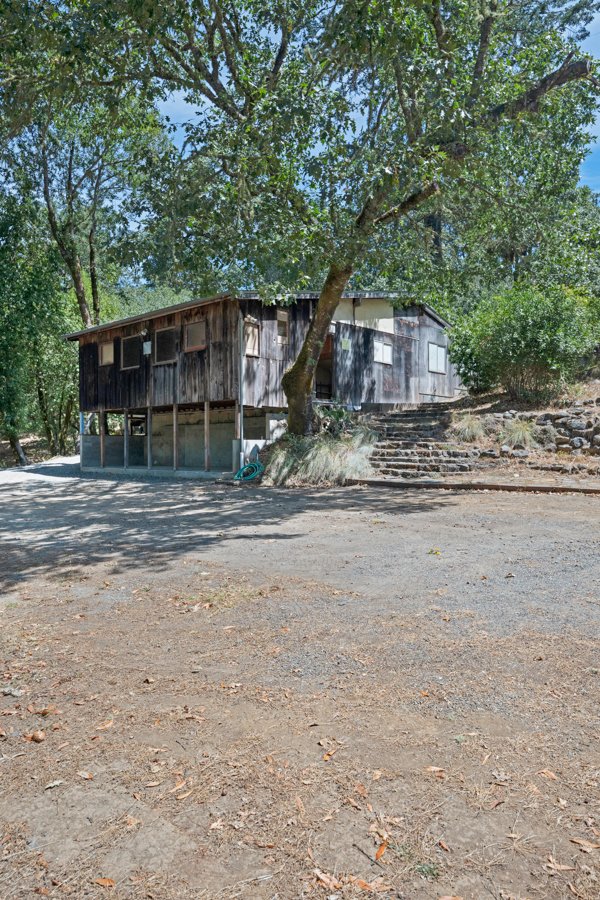 A rustic wooden house on stilts with a shaded area underneath, surrounded by trees and dirt ground, with stone steps leading up to the entrance in a natural setting.