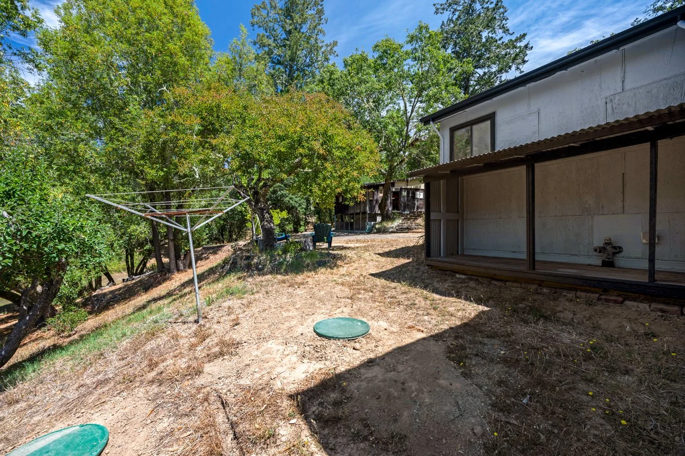 Backyard with trees, a rotary clothesline, a few green chairs, a raised deck, and a water spigot under the overhang.