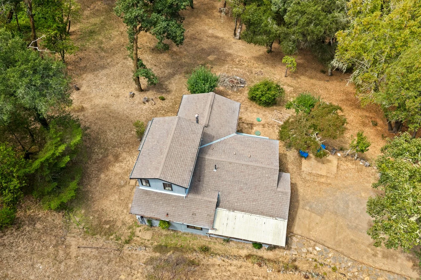 Aerial view of a house in a yard with dry grass, trees around the property, and some outdoor items like a small trampoline and blue chairs.