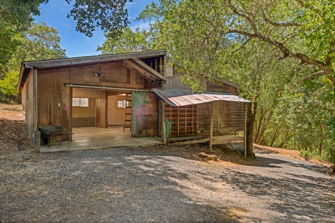 Old wooden barn with open front, surrounded by trees and a gravel driveway.