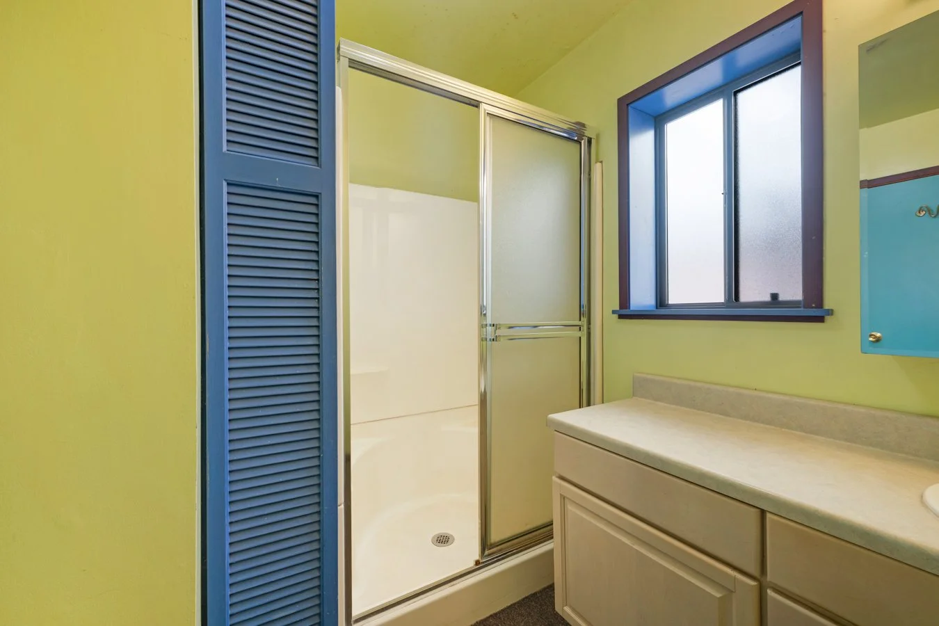 A small bathroom with a glass shower door, a window, a yellow countertop with cabinets, and a blue louvered closet door.