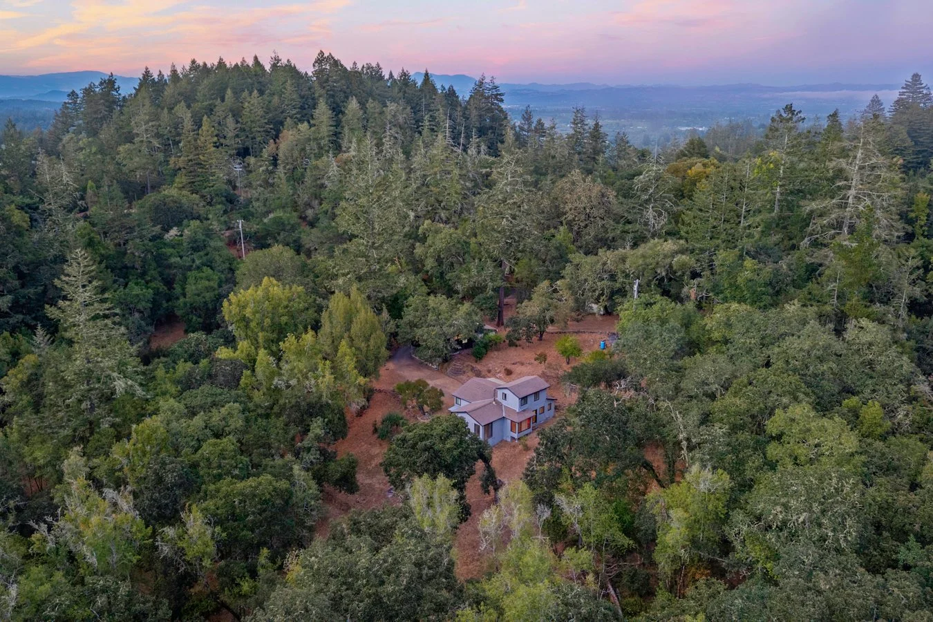 A house surrounded by dense green trees in a hilly forest, with mountains in the background during sunset.
