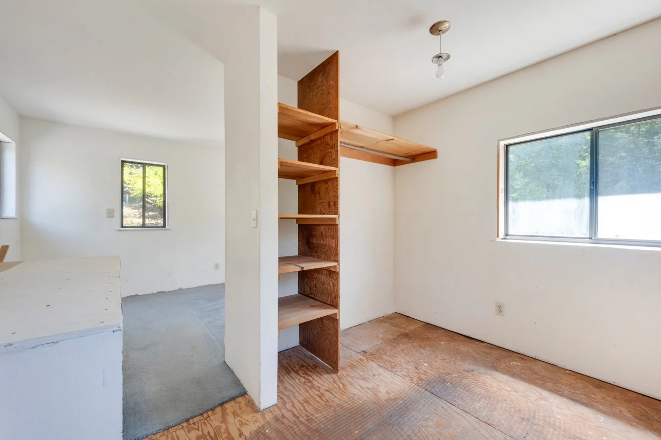 Empty room with white walls, a large window, and wooden shelves built into a partial wall, with some areas showing unfinished flooring.