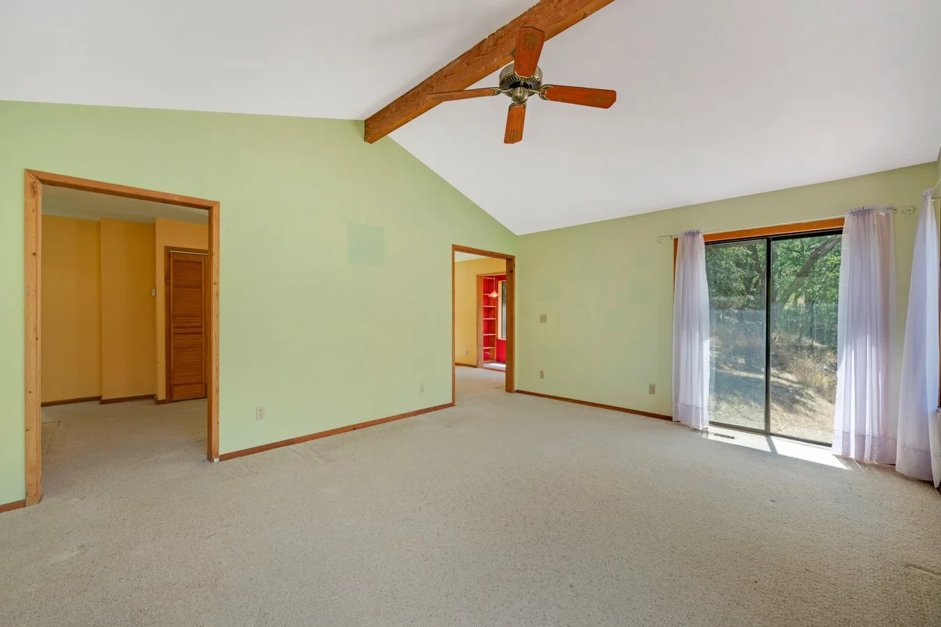 A spacious living room with pale green walls, a vaulted ceiling with exposed wooden beam and ceiling fan, a sliding glass door with sheer white curtains opening to an outdoor area, and doorways leading to other parts of the house.