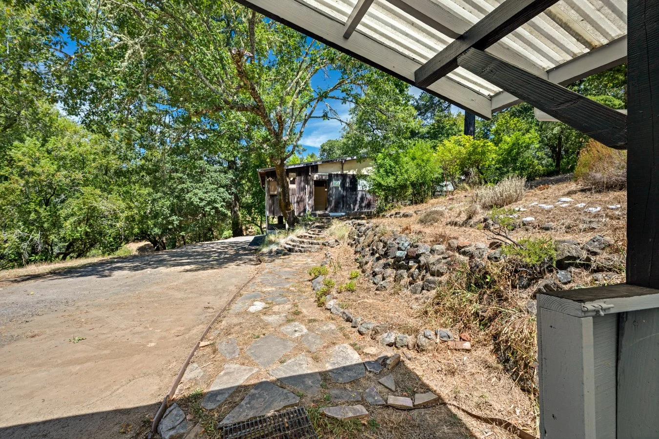 View from a porch or covered area looking towards a dirt pathway, trees, and a small wooden shed on a sunny day.
