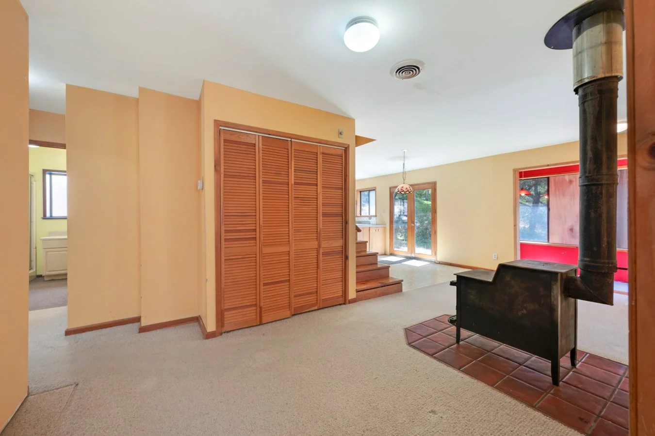 Interior of a house living room with beige walls, wooden cabinetry, a black stove with a pipe, and large windows including a glass door leading outside.