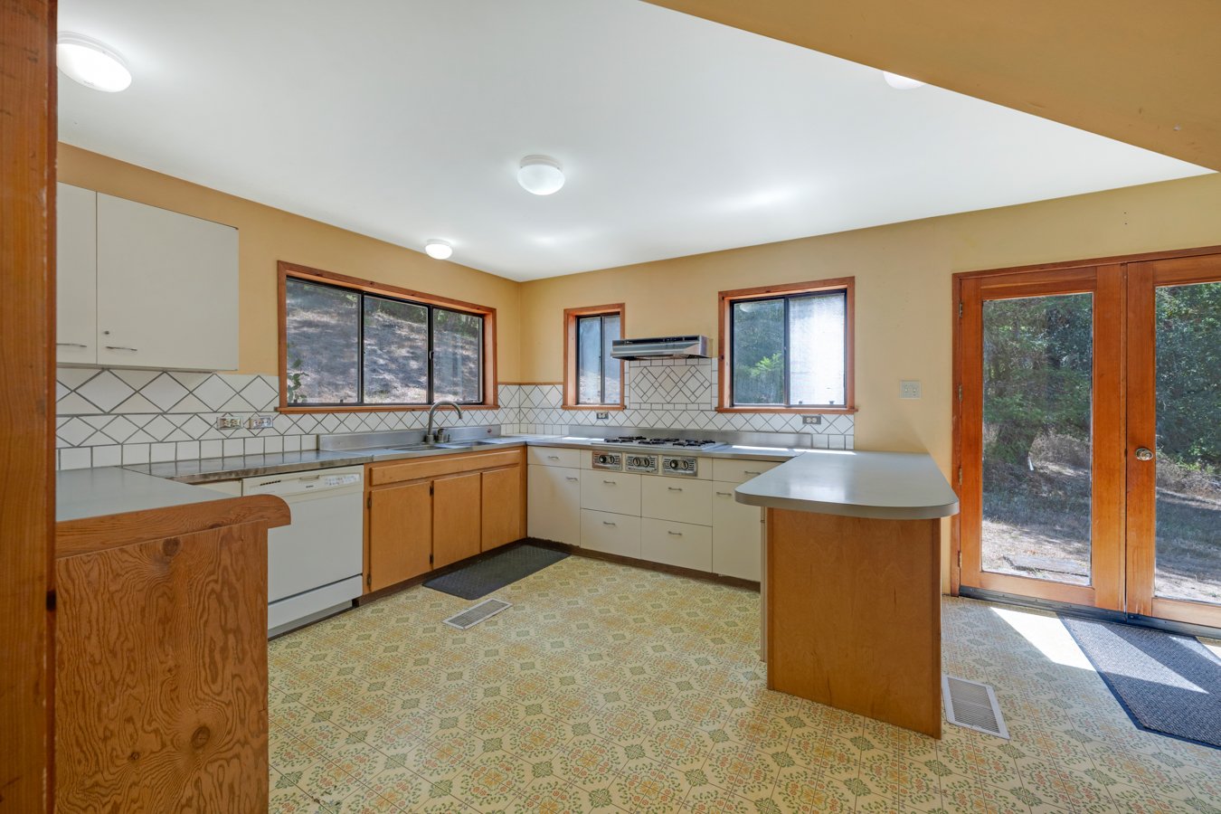 Kitchen with white cabinets, wooden accents, a tiled backsplash, four windows, a sliding glass door, and a yellow patterned linoleum floor.