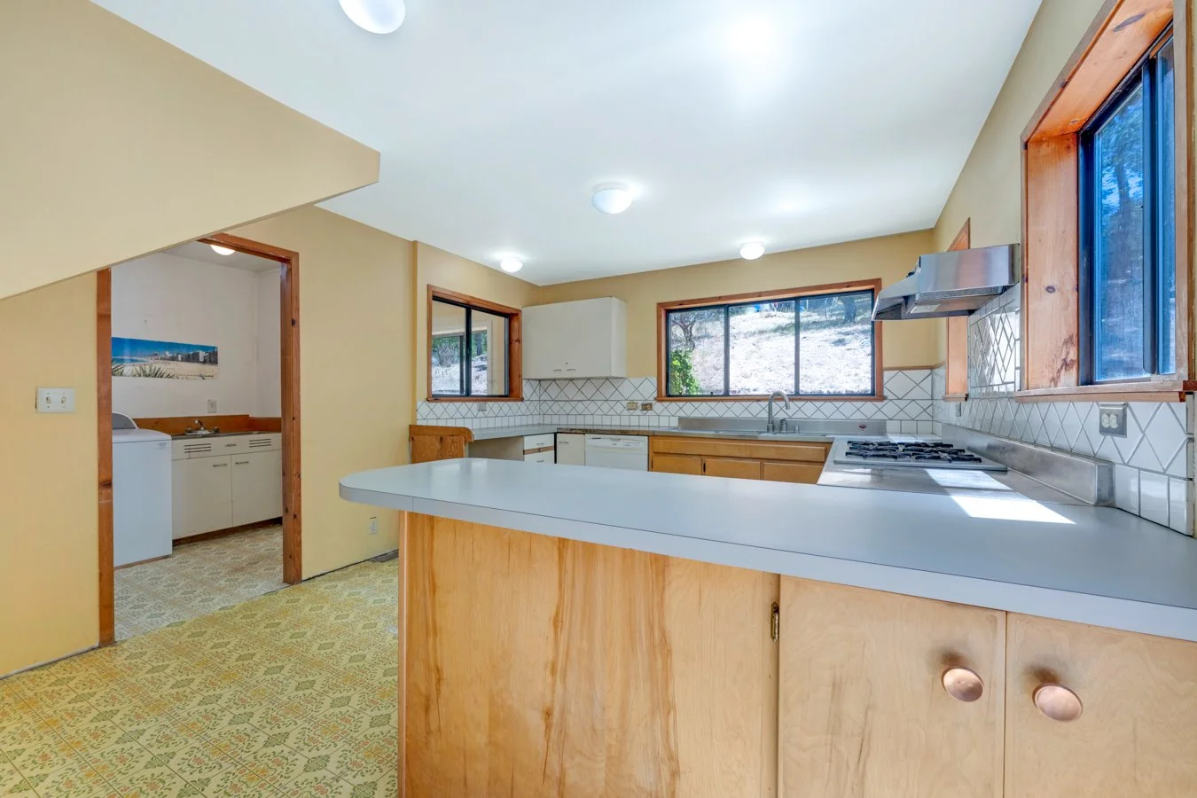 Kitchen with light wood cabinets, white countertops, white tile backsplash, and windows overlooking outdoor scenery, with an adjacent laundry area