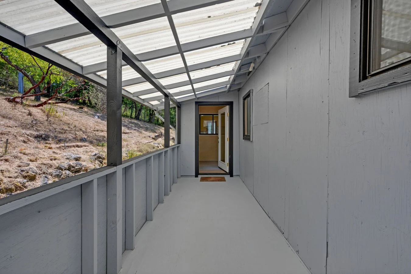 Empty covered porch with white floor and wall, black trim around glass door and window, and a hillside with trees and dry soil outside.