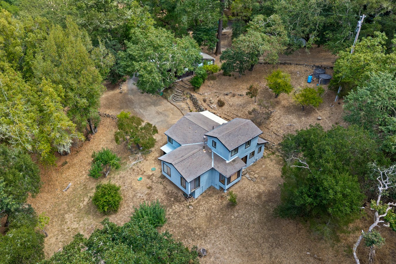 Aerial view of a two-story light blue house with a gray roof, surrounded by trees and a dry yard with some trees and garden features.
