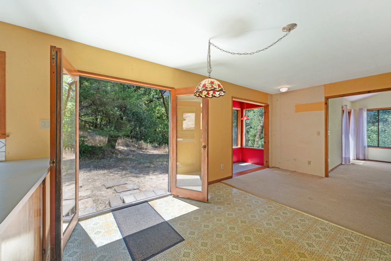 Interior view of a house with an open patio door leading outside, featuring a tiled floor, adjacent to a carpeted room with large windows and curtains, with a hanging lamp and neutral-colored walls.